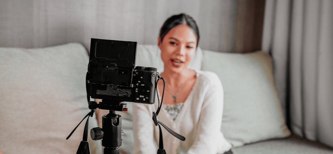 A woman in a sweater films with a DSLR camera on a tripod while seated on a couch, filming a video in a softly lit home setting.