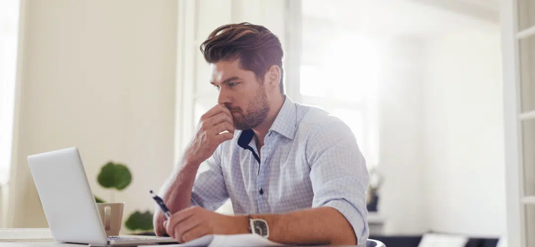 Am I going to get audited by the IRS A worried-looking man in a plaid shirt holds a pen and rests his fist against his mouth, worrying about his tax audits, staring at documents beside a laptop in a bright, airy home office.