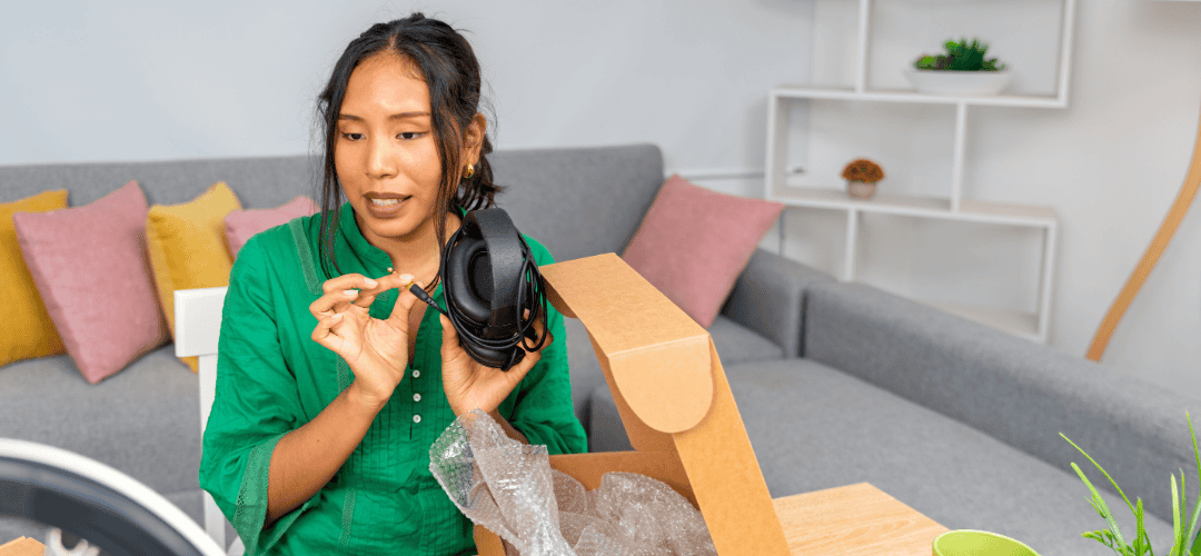 Amazon Influencers A woman in a green shirt unboxes a pair of headphones from a cardboard box, speaking to camera in a bright living room with a ring light visible in the foreground.