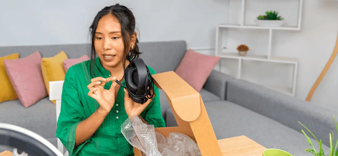 A woman in a green shirt unboxes a pair of headphones from a cardboard box, speaking to camera in a bright living room with a ring light visible in the foreground.