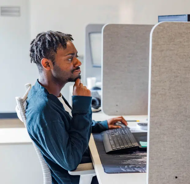 Breon, a Staff Bookkeeper, sits at his desk in a thoughtful pose with his hand on his chin, focused on his computer screen in a bright, modern office with cubicle workstations.