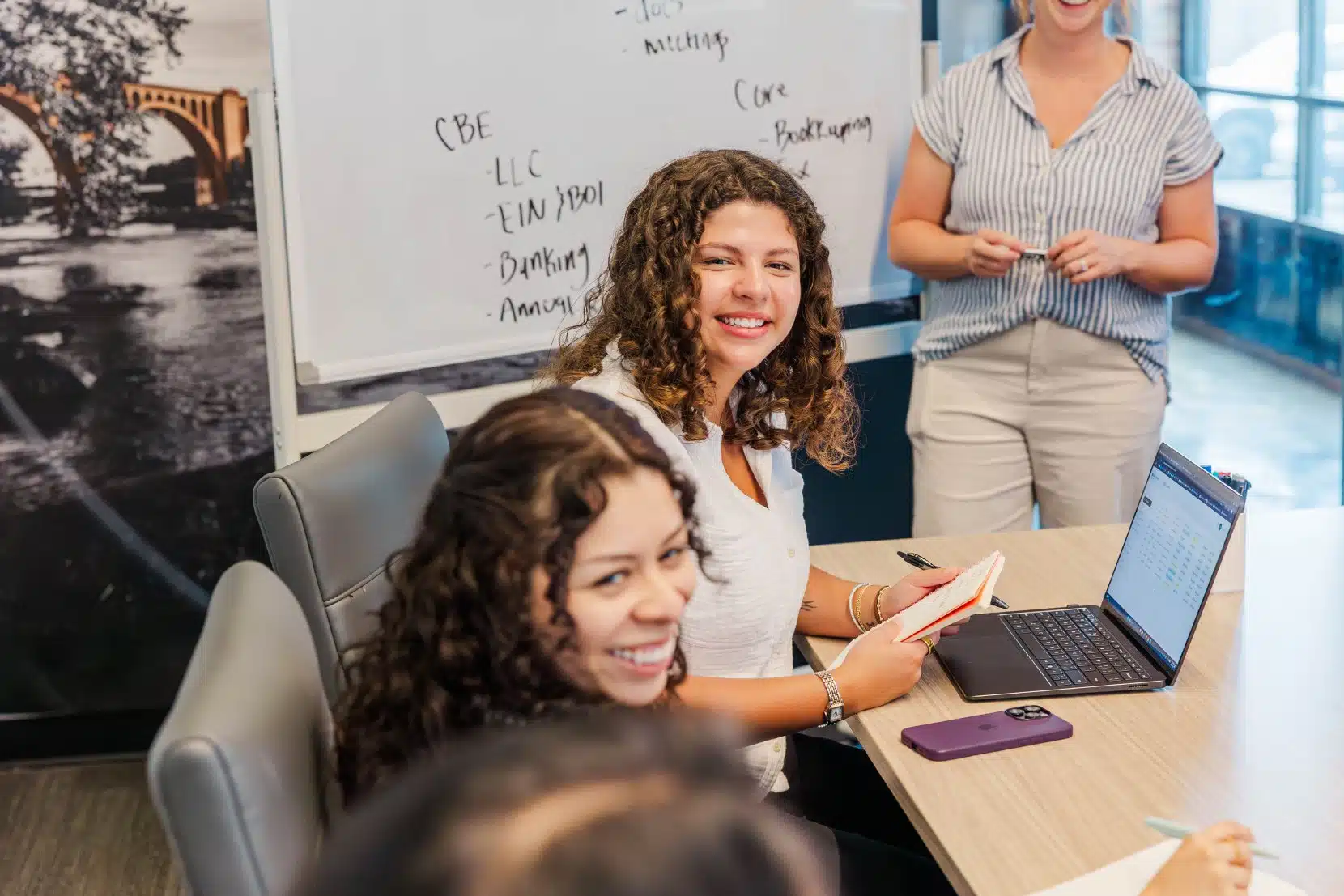 Bri, Tax Associate, seated in the foreground, and Kat, Payroll Specialist, seated behind her, smile during a team meeting in a conference room, with a whiteboard listing business topics visible in the background.