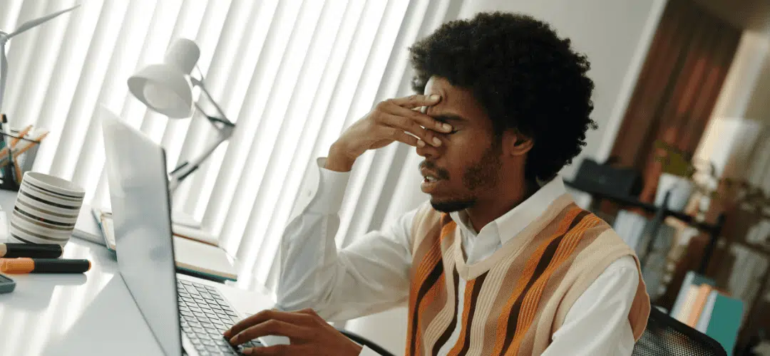 Can Content Creators Get Audited What It Means and How to Avoid It (1) A stressed young man in a striped vest holds his hand over his eyes in frustration while typing on a laptop at a bright home office desk.