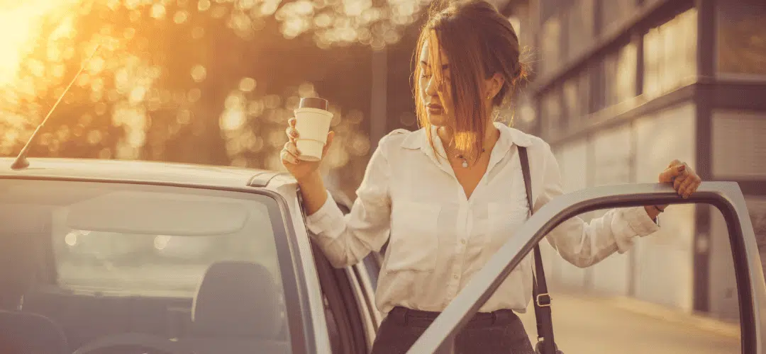 An influencer in a white blouse holds a takeaway coffee cup while stepping out of a car door in warm golden sunlight.