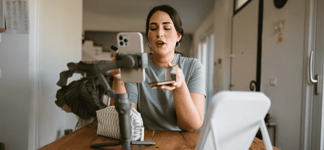 A woman in a light blue t-shirt films herself on a smartphone mounted on a gimbal, holding up a small product while speaking to camera at a home desk.