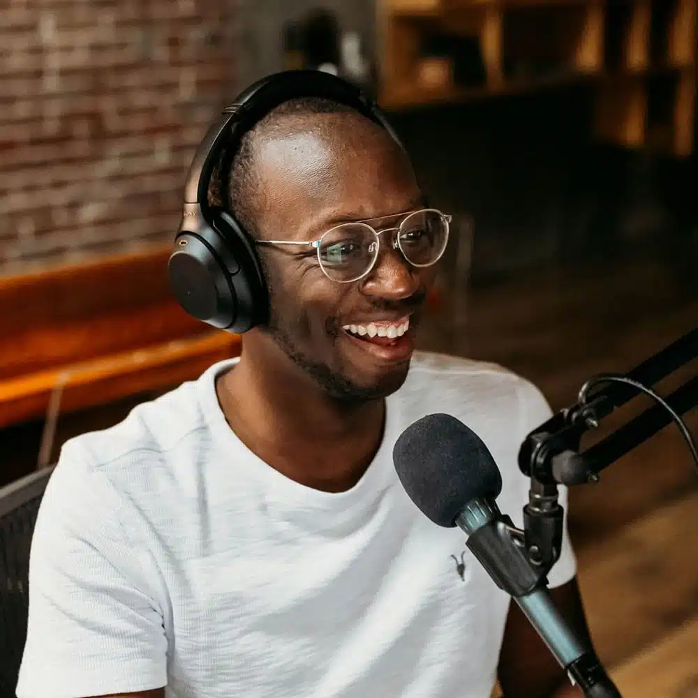 A smiling content creator wearing headphones and glasses speaks into a podcast microphone in a warmly lit studio with exposed brick walls.