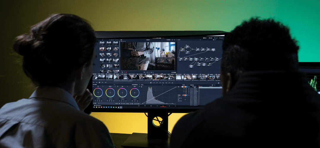 Creators and Contractors Two creators and contractos sit side by side facing a large monitor, reviewing video editing software with timelines, color grading controls, and node-based adjustments displayed on screen.