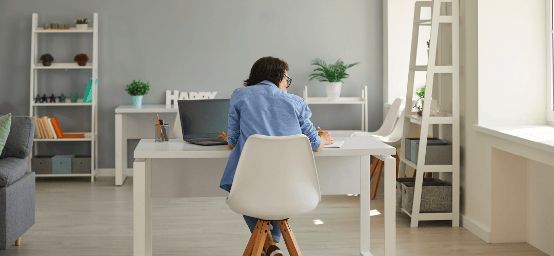Debunking tax myths A woman in a denim shirt sits with her back to the camera, writing notes at a white desk with a laptop in a clean, minimalist home office working on tax myths with white shelving and potted plants.