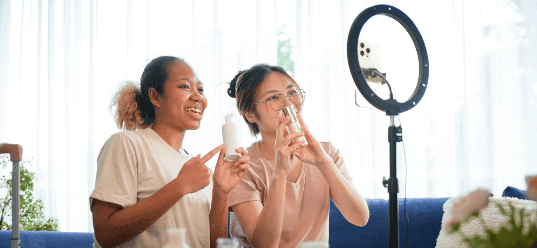 Do I Have to Pay Taxes on PR, Gifts, and Samples as a Content Creator (1) Two young women smile and hold up PR skincare products in front of a ring light with a phone attached, filming a beauty review in a bright home setting.