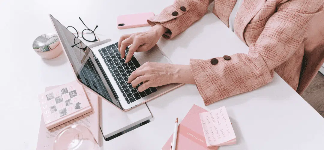 Do You Pay Taxes on Affiliate Marketing A Guide for Content Creators Overhead view of a woman in a pink plaid blazer typing on a laptop at a white desk styled with pink accessories — a notepad with a handwritten to-do list, a pen, glasses, a phone, and a small succulent.