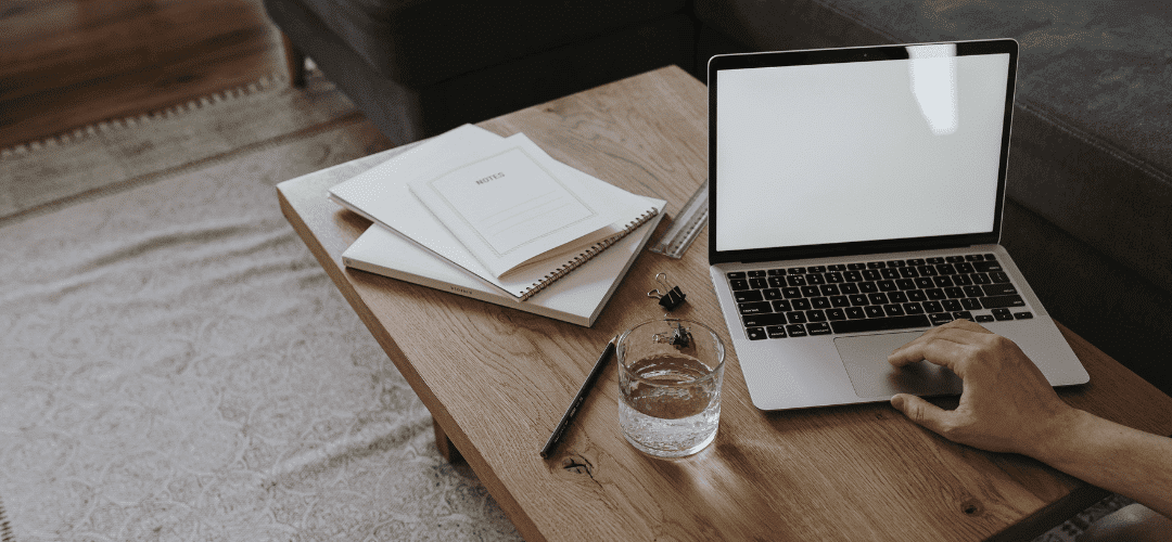 Financial Planning Guide A hand uses a laptop on a rustic wooden coffee table alongside stacked notebooks, a pen, binder clips, and a glass of water, with a couch and rug in the background.