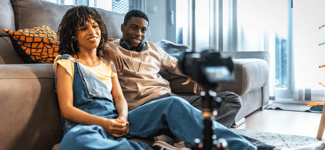 Full Time as a Creator Checklist A young man and woman sit casually on the floor against a couch, smiling at a camera on a tripod as they film content together in a bright living room.
