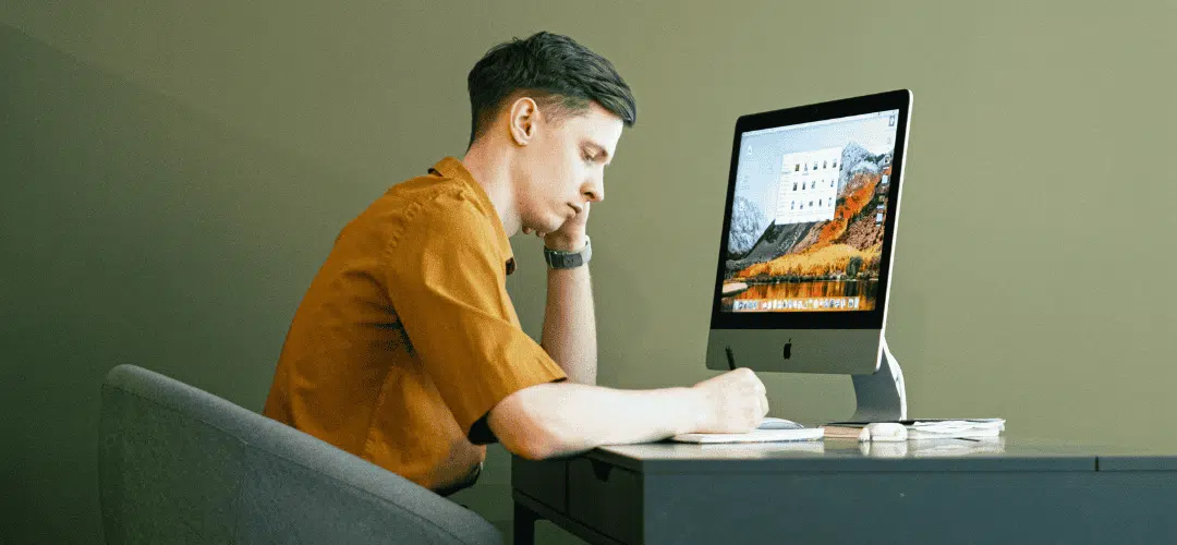 How Content Creators Pay Taxes Simple Guide for New Creators (1) A young man in an orange shirt rests his chin on his hand, staring pensively at an iMac on a minimal desk against an olive green wall.