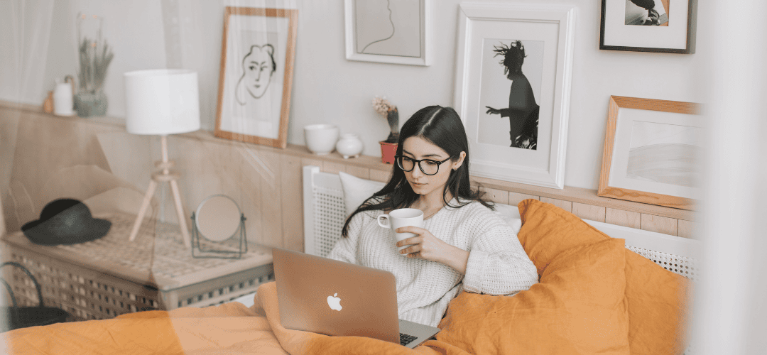 How to Get Financially Organized as a Content Creator A young woman in glasses and a white sweater works on a MacBook in bed, holding a mug, surrounded by yellow pillows and framed wall art in a cozy bedroom.