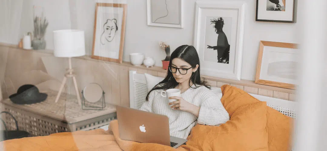 How to Get Financially Organized as a Content Creator A young woman in glasses and a white sweater works on a MacBook in bed, holding a mug, surrounded by yellow pillows and framed wall art in a cozy bedroom.