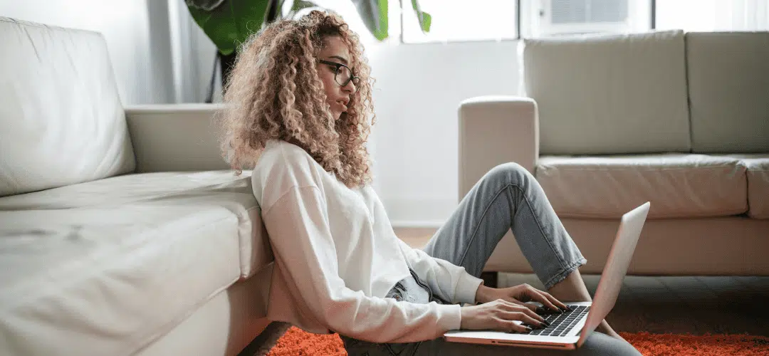 How to Renew your LLC A young woman with curly blonde hair and glasses sits on the floor leaning against a couch, working on a laptop in a bright, casual living room.