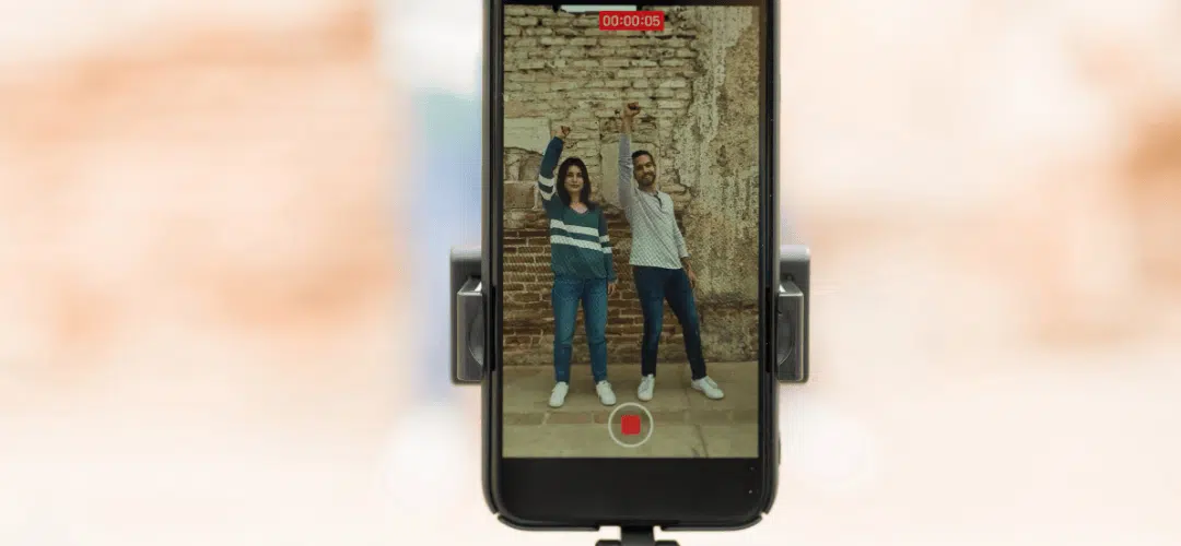 A smartphone mounted on a tripod records two people posing with one arm raised in front of a brick wall, with a red recording indicator visible on the screen.
