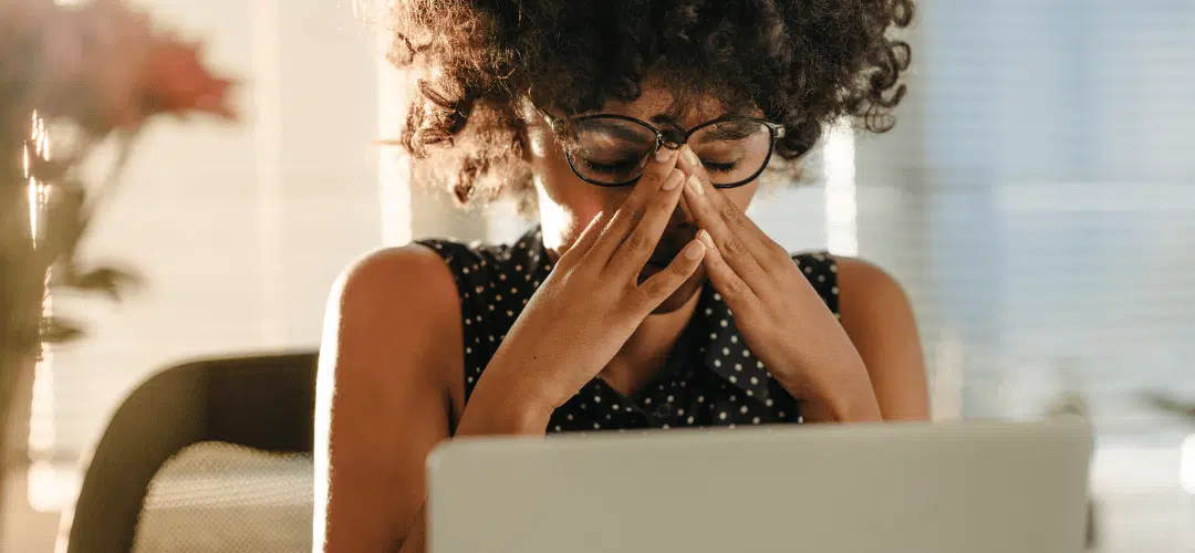 How to file past returns as a content creator A woman in glasses and a polka-dot top presses her hands to her face in exhaustion or stress while filing past tax returns while sitting in front of a laptop in a warmly lit room.