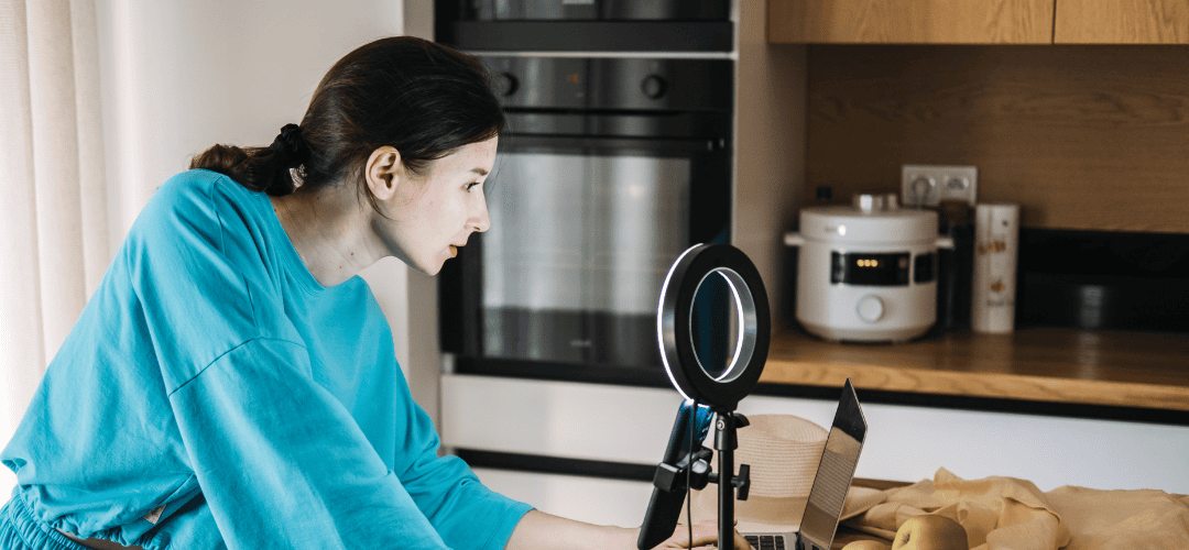 How to file your BOI A woman in a teal top leans toward a ring light and phone setup on a small tripod while working on a laptop, filming content in a home kitchen setting.