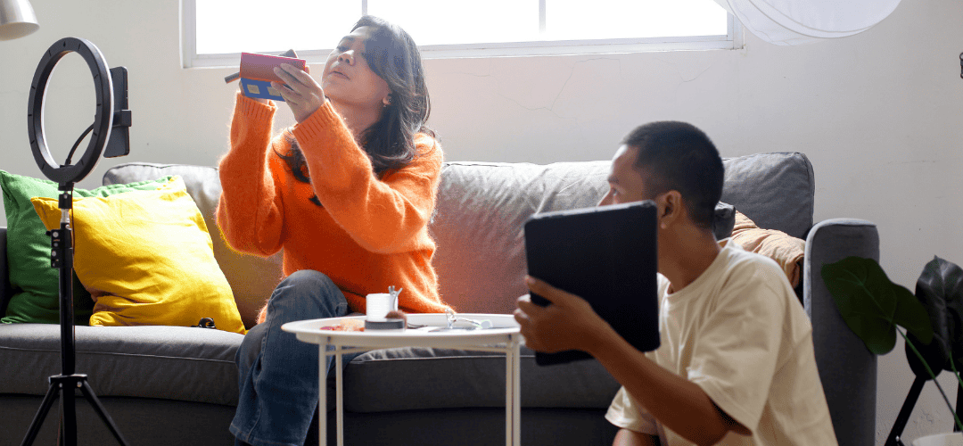 How to get brands to notice you as a content creator A woman in an orange sweater holds up a product on a couch while a man monitors the shot on a tablet, with a ring light set up beside them in a casual home studio, they are trying to get brands to notice them.