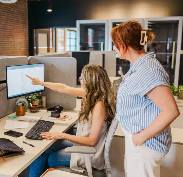 Julia H., Bookkeeping Lead, sits at her desk while Maryn, VP of Operations, stands beside her pointing at the computer monitor, collaborating in a modern office with exposed brick walls and cubicle workstations.