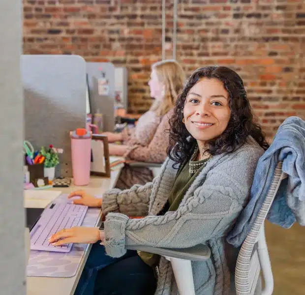Kat, a Payroll Specialist, sits at her desk smiling at the camera while working at her computer with a purple keyboard, in a modern office with exposed brick walls and a colleague working in the background.