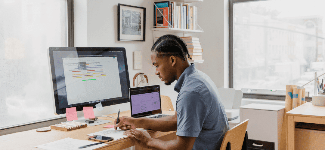 LLC in Wyoming or Florida A man writes notes at a desk with a large monitor and open laptop, surrounded by sticky notes, books, and a printer in a bright, modern workspace.