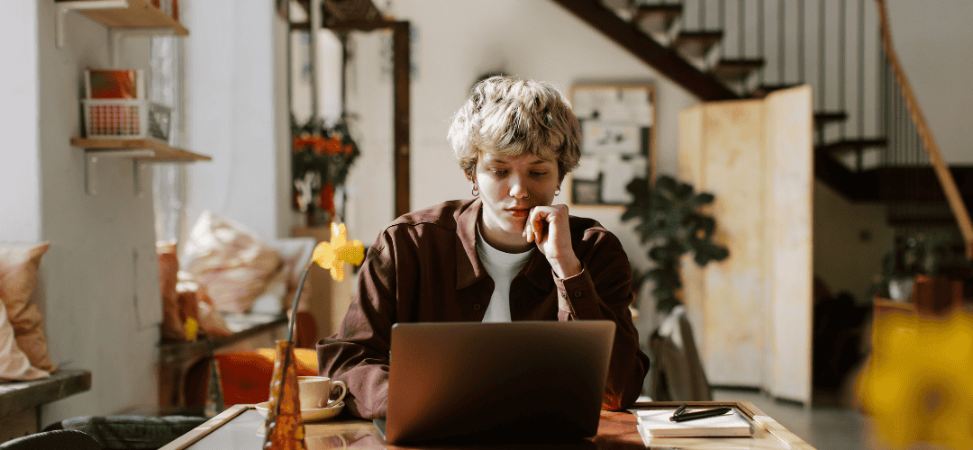 Making Money As a Micro Influencer… Now What A young person with short blonde hair rests their chin on their hand, focused on a laptop in a warm, eclectic home interior with a staircase and cozy decor in the background.