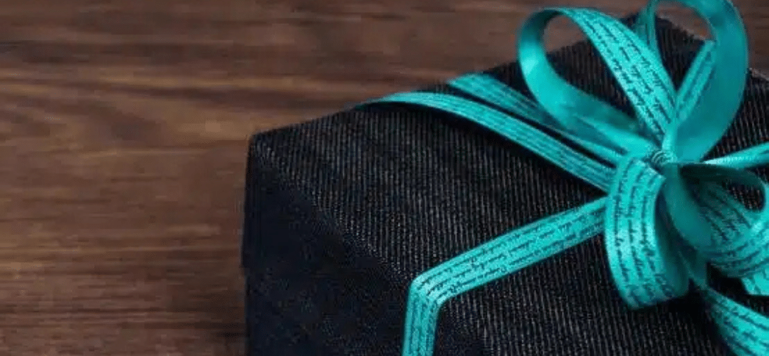 NEW BLOG HEADER Images Less than 200kb (10) A gift box wrapped in dark textured paper and tied with a teal satin ribbon and bow, resting on a wooden surface.