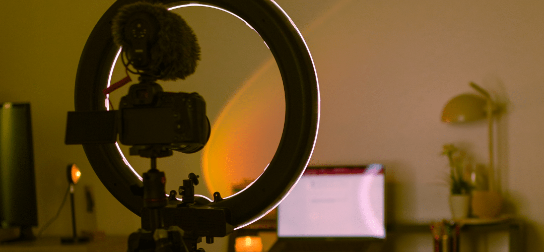 Only Fans Tax blog A ring light with a DSLR camera and shotgun microphone mounted on a tripod, glowing with warm amber light against a dimly lit home studio with a monitor and desk lamp in the background.