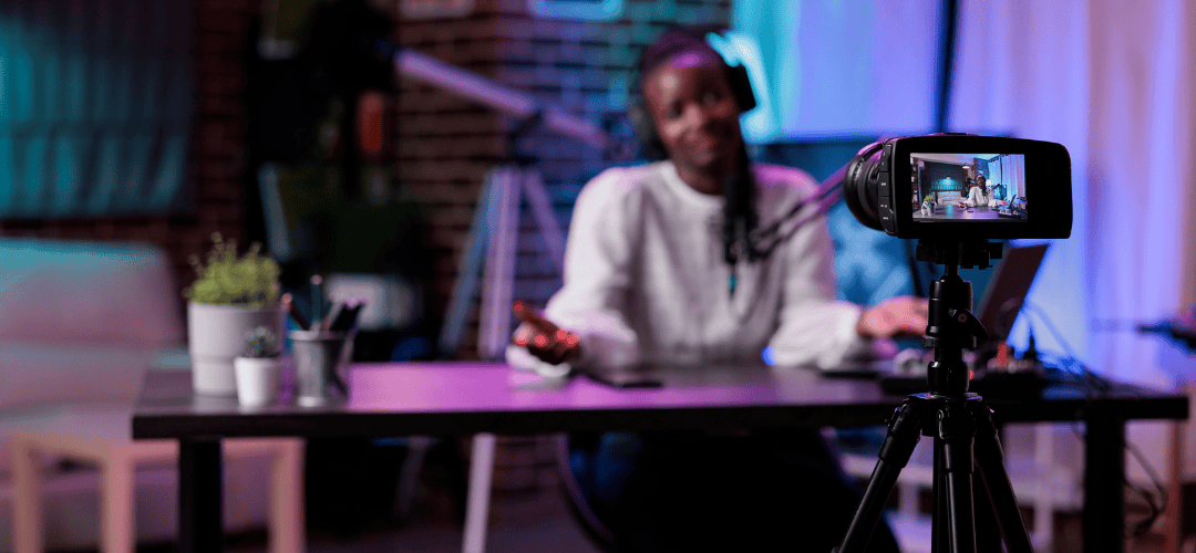 S corp for content creators A smiling man in a white hoodie records himself on a camera with a flip screen in a purple and blue neon-lit studio with a brick wall, desk, and small plant behind him.