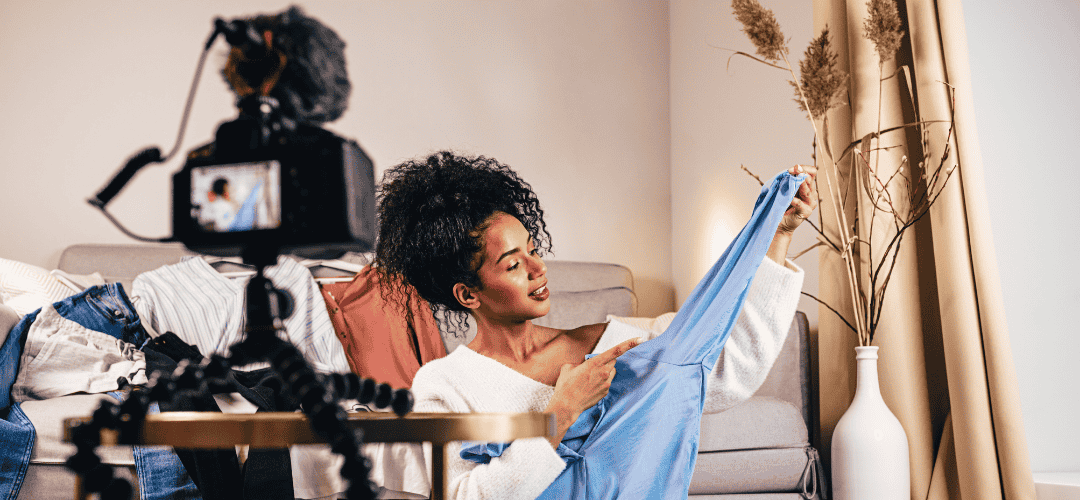 Taxes for Fashion influencers A woman holds up a blue garment and points to it while filming a fashion haul video, with a DSLR camera on a mini tripod and a pile of clothes on a table beside her.