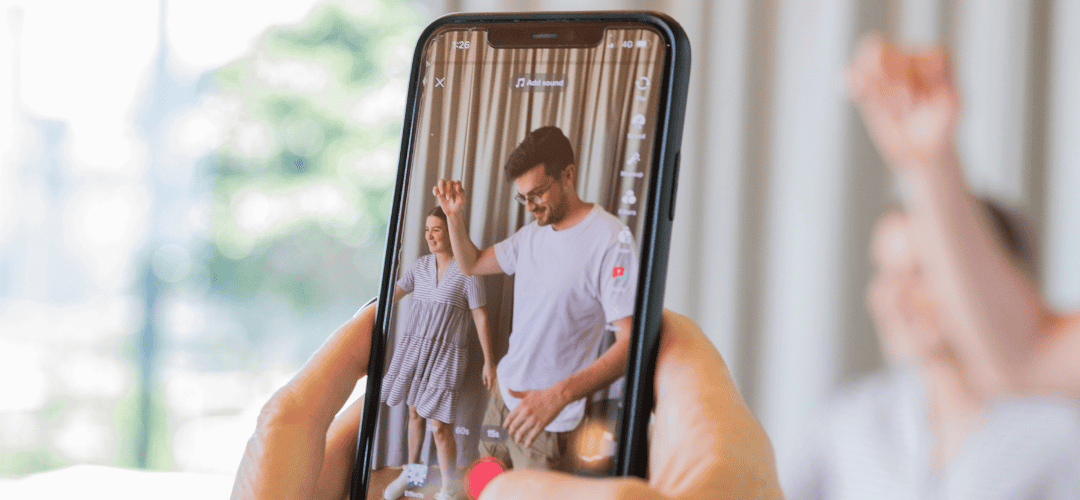 A hand holds up a smartphone recording two people dancing together, with the Tiktok video interface visible on screen and the subjects blurred in the background.