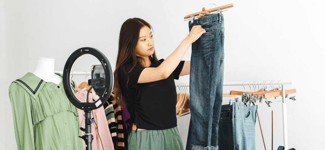 Taxes for WhatNot content creators A WhatNot creator holds up a pair of jeans on a hanger while live-selling in front of a ring light with a phone, surrounded by clothing racks and a mannequin in a clean white studio.