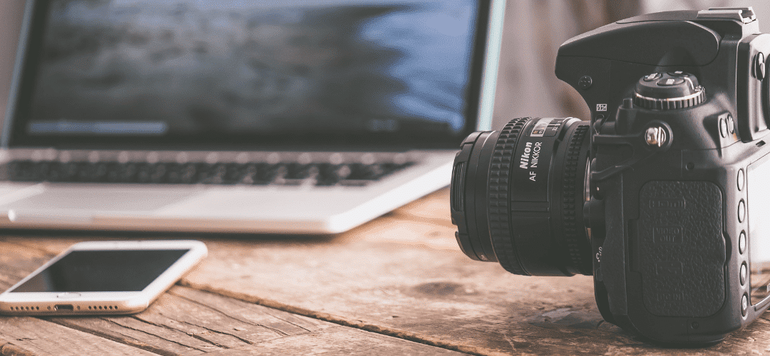 Taxes for onlyfans creators A Nikon DSLR camera sits on a wooden desk beside a smartphone and an open laptop, representing a content creator's equipment setup.