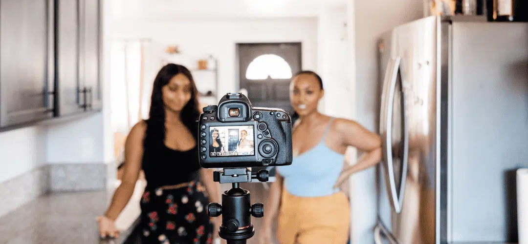 Two women stand in a kitchen filming a Youtube video on a DSLR camera on a tripod, with both of them visible on the camera's rear screen.