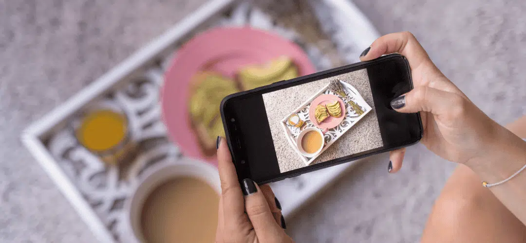 A woman with dark nail polish photographs a styled breakfast tray — with toast, a pink plate, and tea. She is using a smartphone, capturing the flat lay composition on screen.