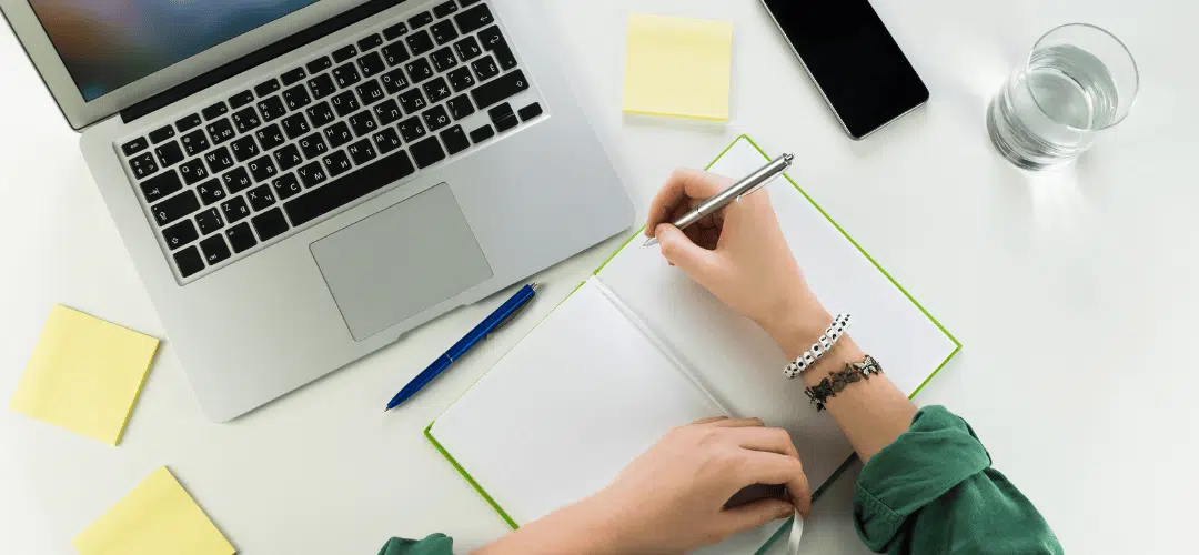 Overhead view of a woman's hands writing in a green notebook beside a laptop, with yellow sticky notes, a pen, phone, and glass of water on a white desk.