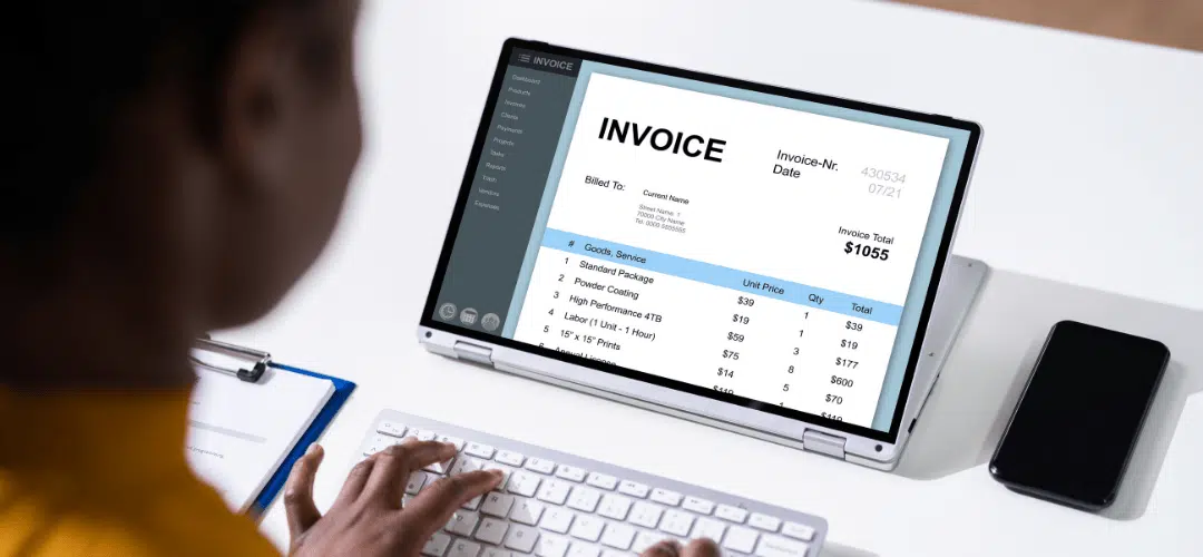 A person types at a keyboard while reviewing a $1,055 invoice displayed on a laptop screen, with a clipboard and phone on the desk beside them.