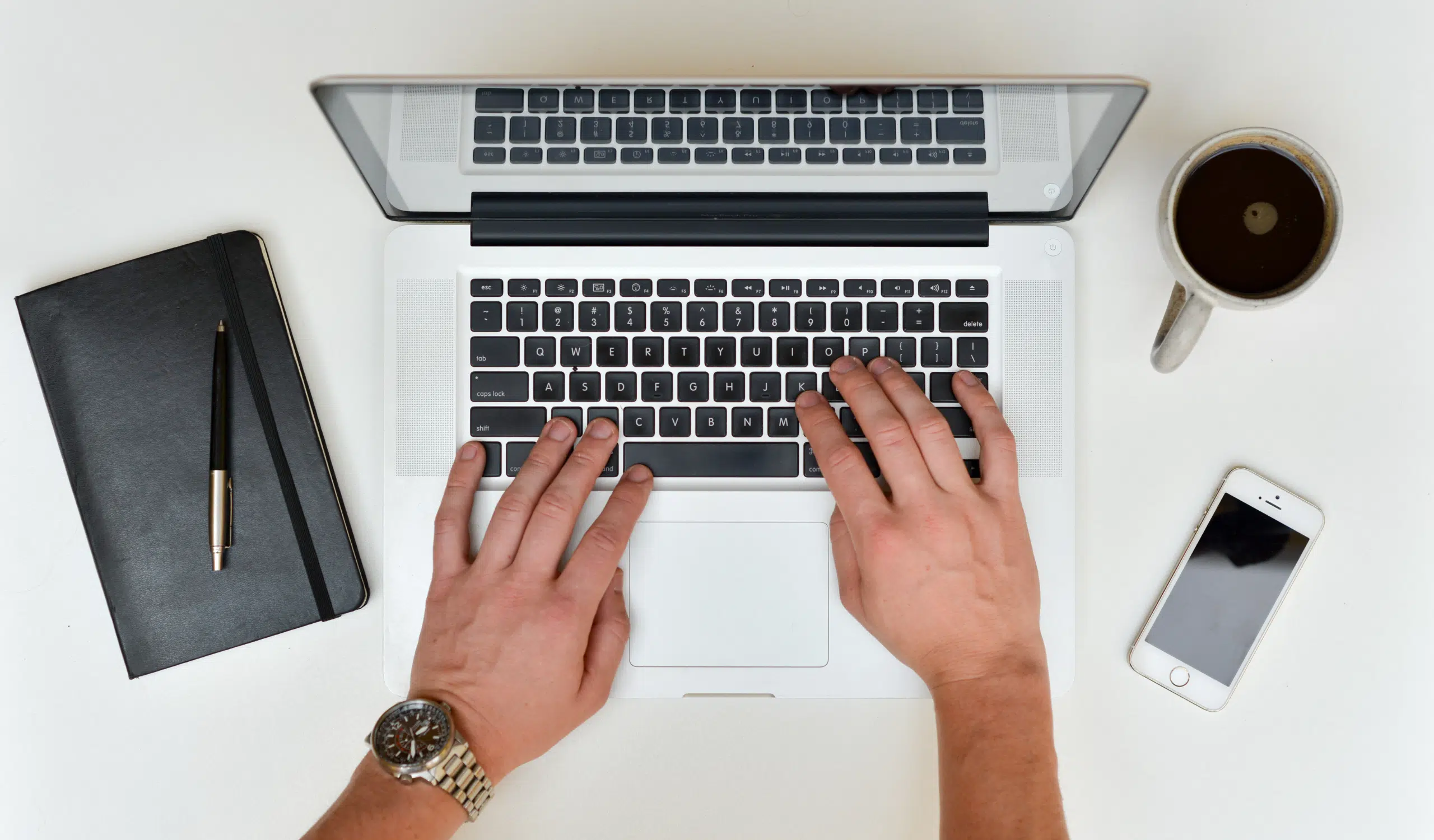Overhead view of a man's hands typing on a laptop trying to get his 1099, with a black notebook and pen, coffee cup, and smartphone arranged on a white desk.