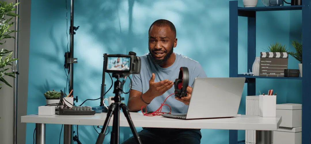 When to pay taxes as a content creator A man holds up a pair of headphones and gestures while recording a tech review video, with a DSLR camera on a tripod, laptop, studio light, and clapperboard visible in his blue-walled home studio.