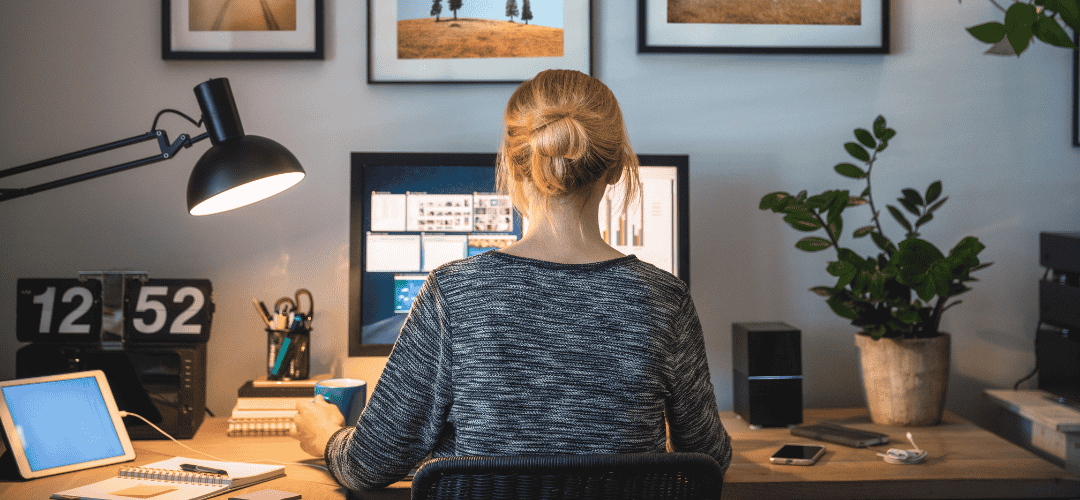bookkeping for content creators A woman with her hair up sits with her back to the camera, working at a desktop monitor in a warmly lit home office with a desk lamp, flip clock, plant, tablet, and landscape prints on the wall.