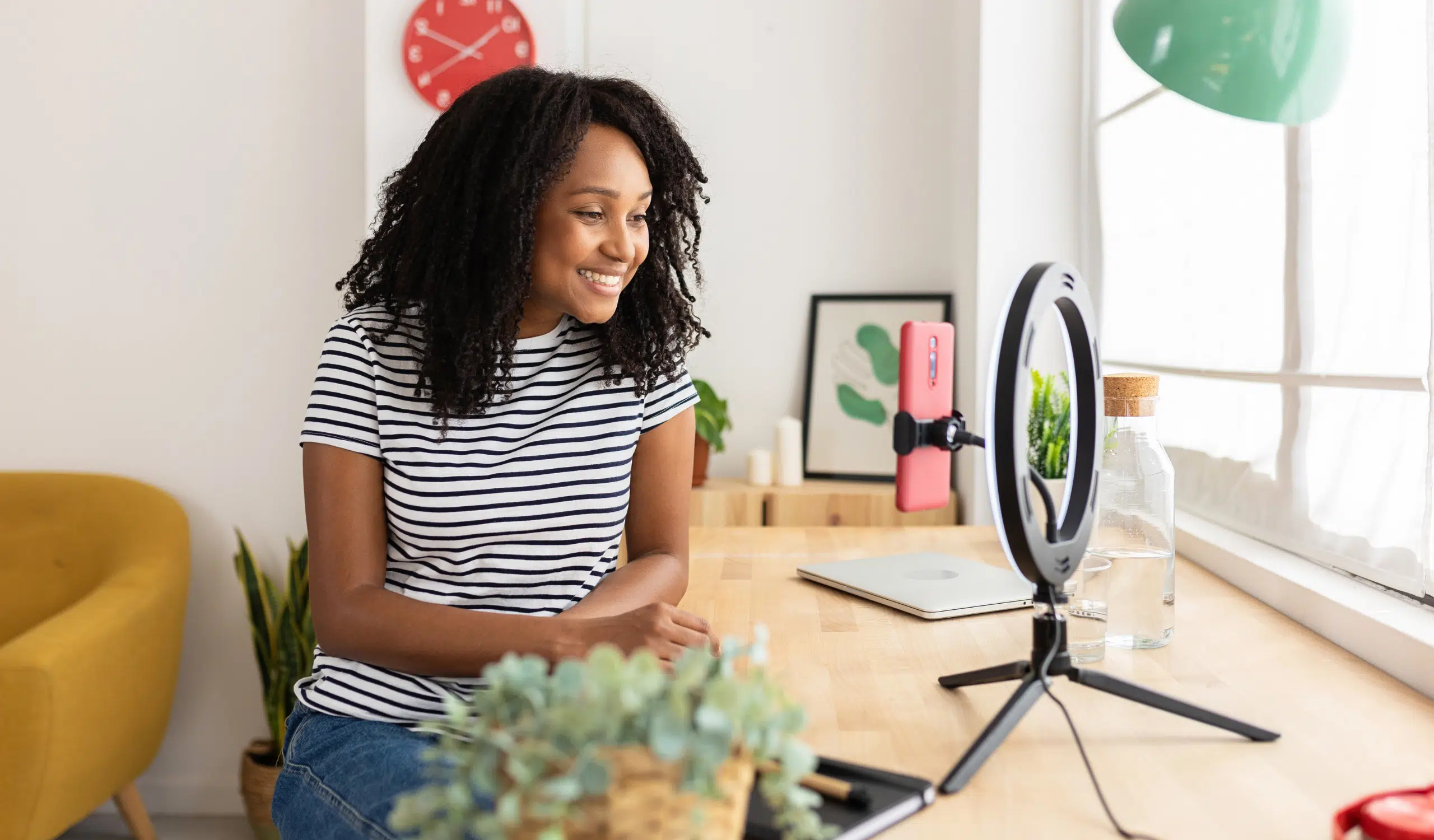 brand deals (2) A smiling woman in a striped t-shirt records content at a bright home desk, with a phone mounted on a ring light, a laptop, and a red clock on the wall behind her.