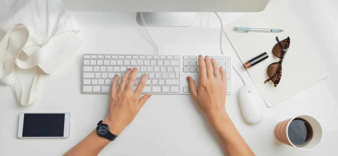 common tax filing mistakes as an influencer Overhead view of a woman's hands typing on an Apple keyboard, filing her taxes with sunglasses, lipstick, a pen, phone, mouse, and coffee cup arranged on a clean white desk.