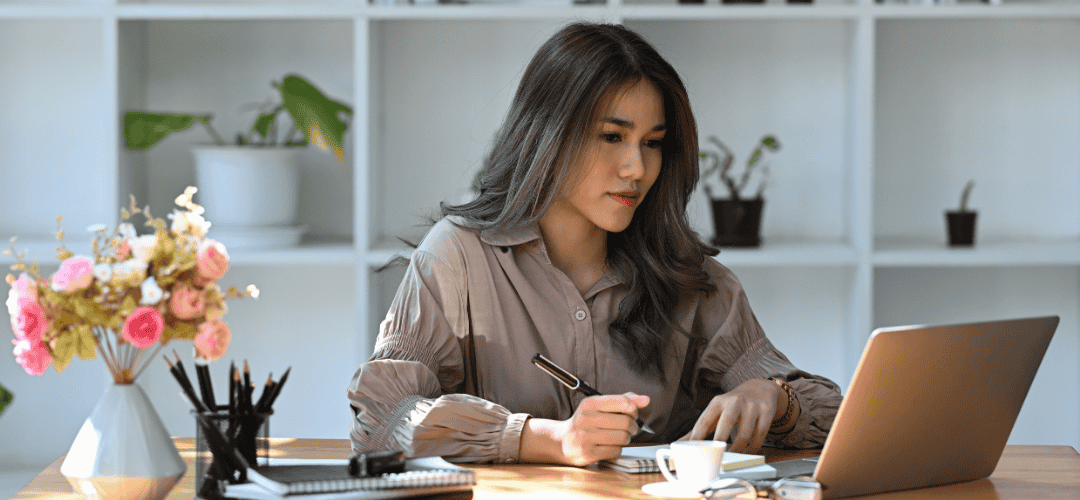 content creator tax forms (2) A woman in a taupe blouse writes notes at a desk while looking at a laptop, with a floral arrangement, pencil holder, and coffee cup nearby and white shelving with plants in the background.