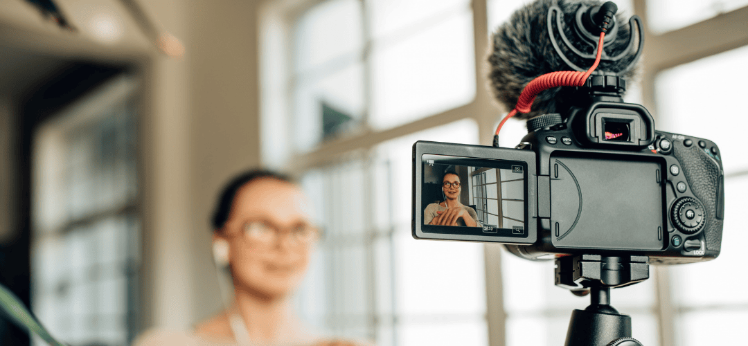 content creator tax forms A DSLR camera with a shotgun microphone on a tripod shows a woman in glasses on its flip screen as she records herself in a bright, windowed room.
