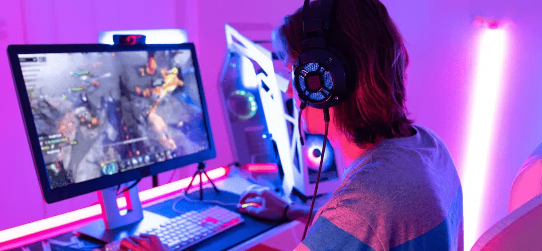 A person wearing large gaming headphones plays an action game on a monitor at an RGB-lit gaming desk with a white PC tower, bathed in pink and purple neon light.