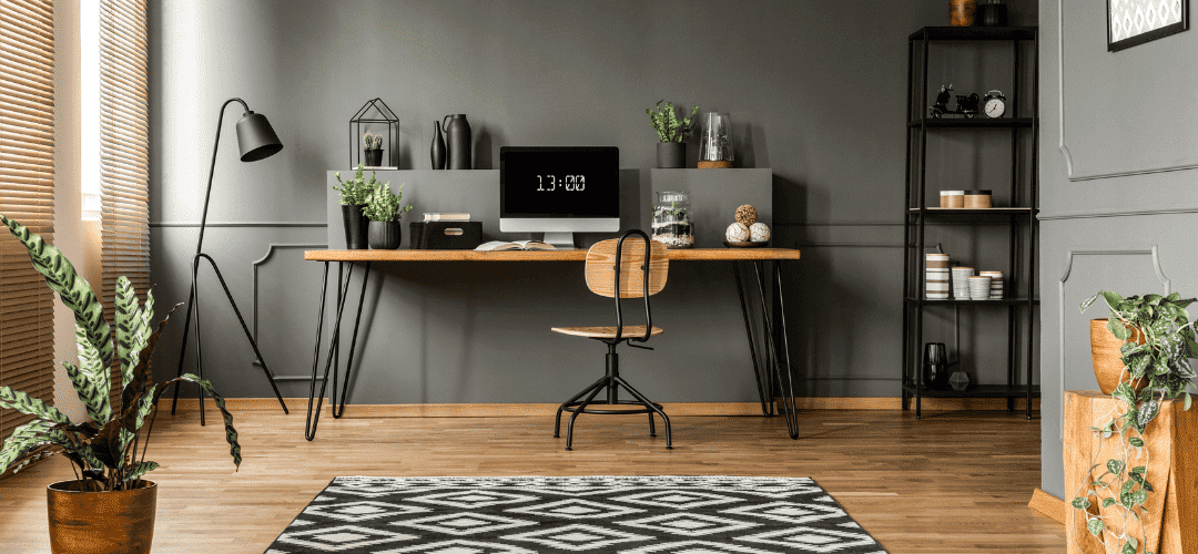 home office deductions A stylish home office with a wooden hairpin-leg desk, iMac, wooden swivel chair, black floor lamp, metal shelving, potted plants, and a geometric rug against a dark gray wall.