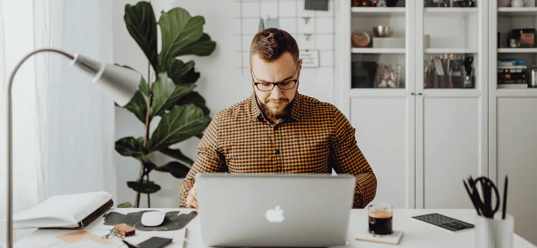 A bearded man in glasses and a plaid shirt works focused on a MacBook at a tidy white desk, with a desk lamp, coffee, and organized shelving in the background.