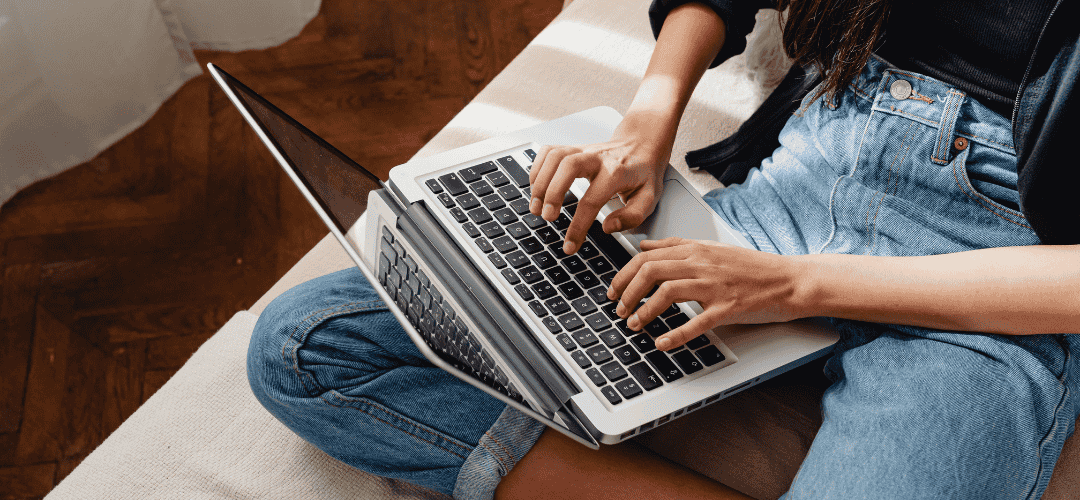 tax deductions 101 (2) Overhead view of a woman in jeans sitting cross-legged on the floor, typing on a laptop balanced on her knees over a white rug and hardwood floor.